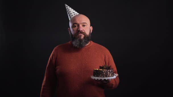 Un hombre barbudo sobre un fondo negro vestido con una gorra festiva y sosteniendo un pastel en sus manos una serpentina cae sobre él — Vídeos de Stock