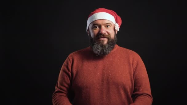 Um homem barbudo em um fundo preto vestido com um chapéu de Papai Noel segura uma caixa em suas mãos e se alegra — Vídeo de Stock