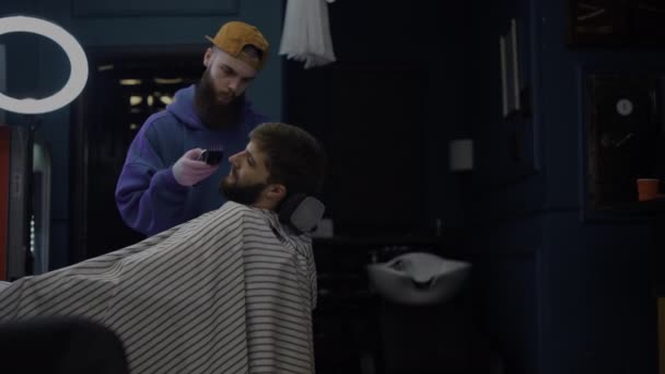 Um homem barbudo corta a barba numa barbearia. Barbeiro funciona como um trimer em um salão — Vídeo de Stock