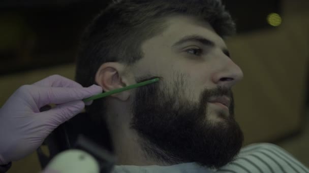 Um homem barbudo corta a barba numa barbearia. Fechar o rosto e as mãos de um barbeiro — Vídeo de Stock