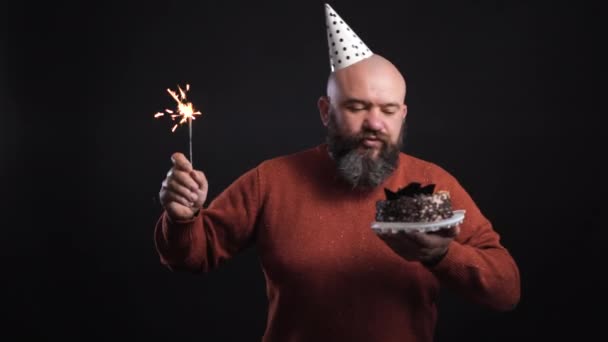 Γενειοφόρος άνδρας σε μαύρο φόντο ντυμένος με εορταστικό καπέλο και κρατώντας μια τούρτα και ένα λαμπόγυαλο, χορεύει — Αρχείο Βίντεο