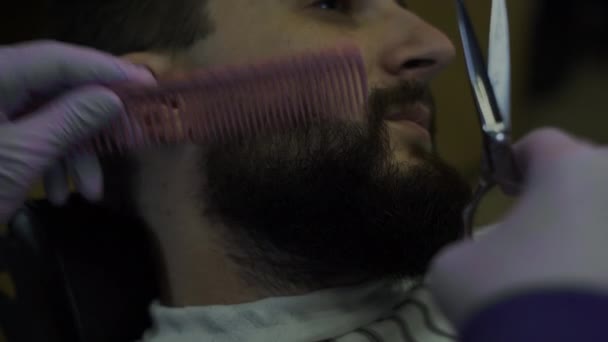 Бородатий чоловік обертає бороду в перукарні. крупним планом перукар розчісує волосся і ріже ножицями — стокове відео