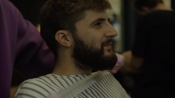 Бородатый мужчина сидит в парикмахерской. Fix the cape for the client — стоковое видео