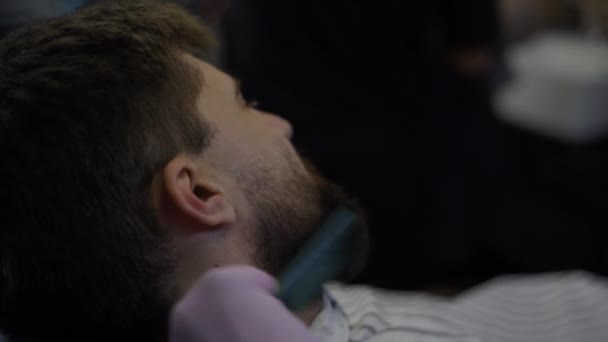Um homem barbudo corta a barba numa barbearia. Close-up de mãos de cabeleireiro trabalhando com trimer — Vídeo de Stock