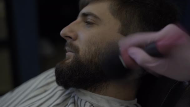 Un hombre barbudo en una barbería, un peluquero con un cepillo quita los restos de pelo de su cara — Vídeo de stock