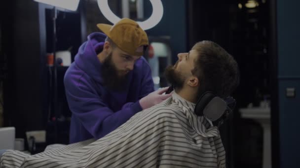 Un hombre barbudo se sienta en un sillón, un peluquero con una cortadora de pelo hace una corrección de barba — Vídeos de Stock