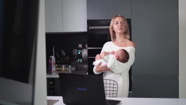 Młoda piękna kobieta stoi w kuchni w pobliżu laptopa i trzyma małe dziecko w ramionach. — Wideo stockowe