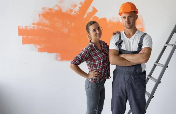 young stylish Caucasian man dressed in overalls and orange safety helmet. The woman is standing nearby.