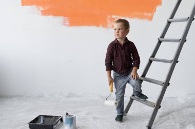 Ciddi bakışları olan genç bir çocuk merdivenlerin yakınındaki bir odada duruyor. Elinde boya fırçası tutuyor..