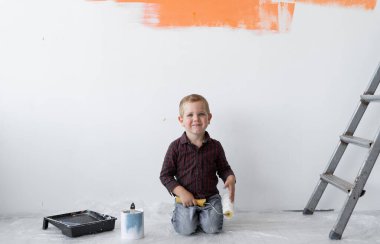 Gülümseyen genç bir çocuk merdivenlerin yanındaki bir odada oturuyor. Elinde boya fırçası tutuyor. Ev yenileme konsepti.