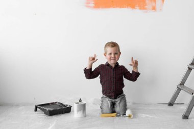 Gülümseyen genç bir çocuk merdivenlerin yanındaki bir odada oturuyor. İşaret parmağı havadayken boya silindirini elinde tutuyor. Ev yenileme konsepti.