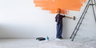 Tulum giymiş ve koruyucu miğfer takmış bir çocuk duvarın yanında durup onu fırçayla boyuyor. Ev yenileme konsepti.