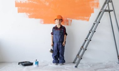 Tulum giymiş ve koruyucu kasklı bir çocuk elinde fırçayla duvarın yanında duruyor. Ev yenileme konsepti.