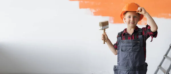 A boy dressed in overalls stands in a room holding a paintbrush in his hands, takes off a protective helmet from his head. Home renovation concept. — Stock Photo, Image