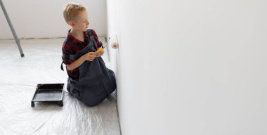 Tulum giymiş bir çocuk odada elinde boya ile bir silindir tutuyor ve duvarı boyuyor. Ev yenileme konsepti.