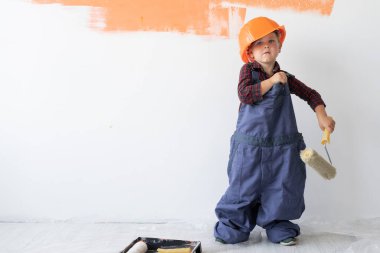 Kafasında turuncu bir miğfer olan küçük bir çocuk duvarın yanında duruyor ve büyük bir tulumu düzeltiyor..