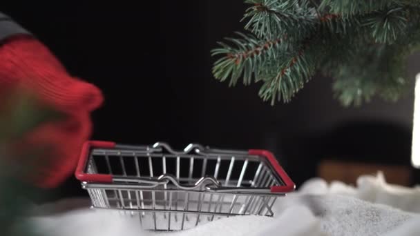 Op een stapel sneeuw onder de boom, een hand in een handschoen zet een boodschappenmandje en een rode doos met een geschenk wordt er in gegooid — Stockvideo