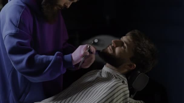 한 남자가 바베큐 의자에 앉아 있습니다. 헤어 드레서는 면도칼로 고객의 면도칼을 깎는다 — 비디오