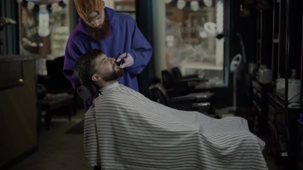 Un hombre se sienta en un sillón, un barbero se corta la barba. Concepto de pequeña empresa — Vídeo de stock