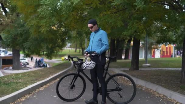En ung man står nära cykeln, tar av sig hjälmen från huvudet och hänger den på styret. aktivt livsstilsbegrepp — Stockvideo