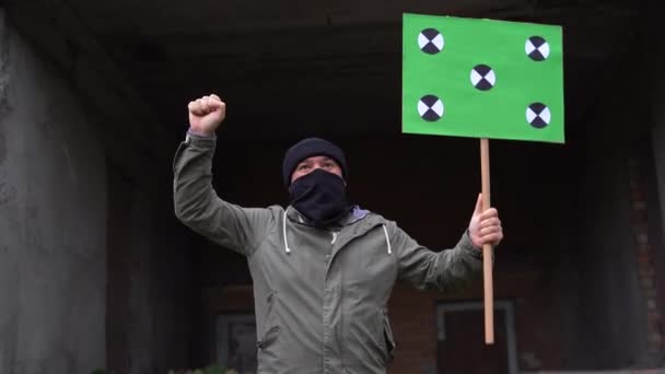 黒い顔のマスクをした男がポスターを手に持っている。スペーストラッキングポイントをコピーします。空白の緑のスクリーンボード. — ストック動画