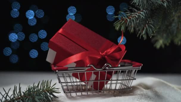 Czerwone pudełko ze wstążką leży pod choinką w koszyku na zakupy. Koncepcja prezenty Boże Narodzenie — Wideo stockowe