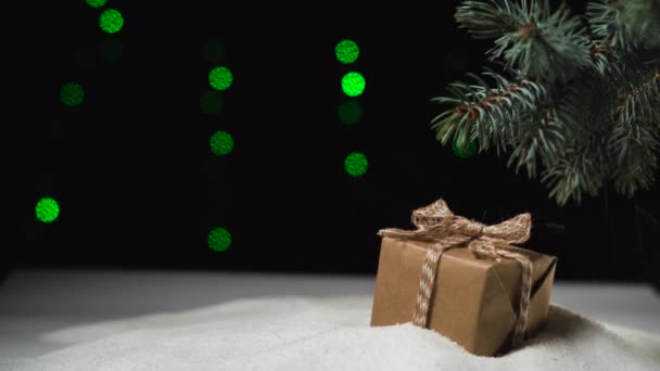 En la nieve bajo el árbol se encuentra una caja con un arco, papel artesanal. nieve fina está cayendo. Luces de Navidad — Vídeo de stock