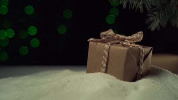 A hóban a fa alatt fekszik egy doboz egy íj, kézműves papírt. Karácsonyi fények — Stock videók