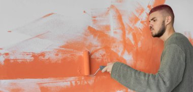 Genç bir adam bir silindirle parlak turuncuya duvar boyuyor. İnşaat bitirme işleri.