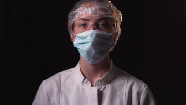 Giovane donna medico vestito con un cappotto bianco, una maschera medica sul viso e occhiali di sicurezza. Sta su uno sfondo nero e guarda tranquillamente la fotocamera — Video Stock