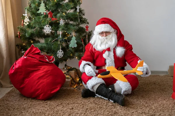 Santa Claus duduk di lantai dekat pohon Natal, membuka tas dan mengambil hadiah pesawat mainan. Dia lelah dan duduk untuk beristirahat. — Stok Foto