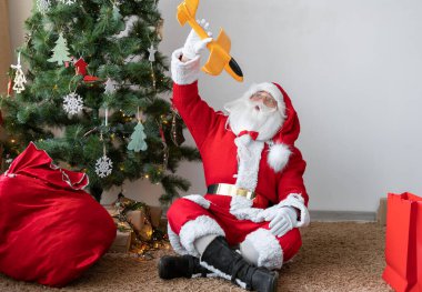 Noel Baba, Noel ağacının yanında, içinde hediyeler olan bir çantanın yanında yerde oturuyor. Oyuncak bir uçakla oynuyor..
