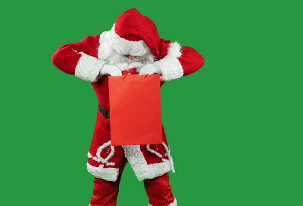 Der Weihnachtsmann auf grünem Grund hält eine rote Einkaufssuppe in der Hand und schaut hinein. Kopierraum. — Stockfoto