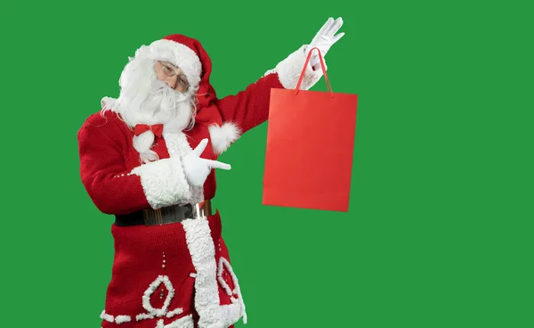 Kerstman op een groene achtergrond houdt een rode boodschappentas in zijn handen tilt en wijst er met zijn vinger naar. Kopieerruimte. — Stockfoto