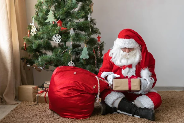 santa claus sits on the floor near the christmas tree untied his bag and took out a gift. box with red ribbon in hands.