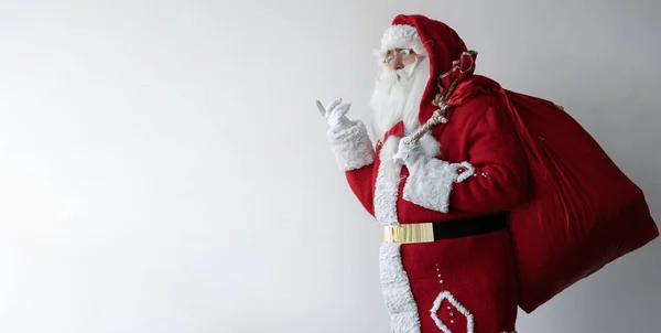 Santa claus está de pie sobre blanco, sosteniendo una gran bolsa detrás de su espalda y pensando en algo. —  Fotos de Stock