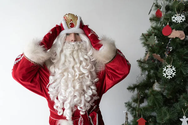Ο Άγιος Νικόλαος στέκεται δίπλα στο δέντρο της Πρωτοχρονιάς στο δωμάτιο και βάζει ένα καπέλο στο κεφάλι του. Φοράει κόκκινο κοστούμι και γάντια. Κοντινό πλάνο. — Φωτογραφία Αρχείου