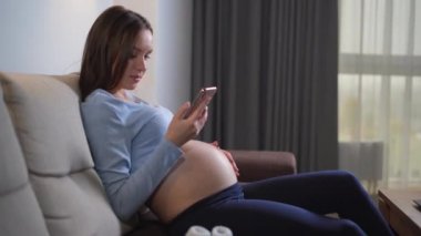 Genç hamile bir kadın evinde, kanepede oturuyor. Telefonda bir mesaj yazıyor..