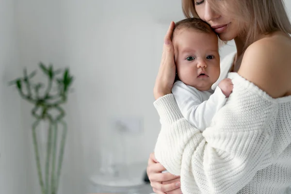 Mamá sostiene a un bebé recién nacido en sus brazos y presiona su cabeza contra su pecho. Mujer y niño en el dormitorio sobre un fondo blanco. Maternidad y concepto infantil. — Foto de Stock