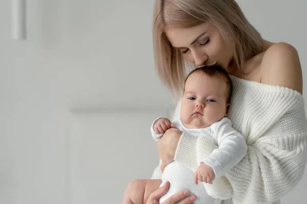 Una mujer con un bebé recién nacido en sus brazos besa la cabeza de sus hijas. Ropa confeccionada en algodón blanco de calidad. Lugar para su texto. — Foto de Stock