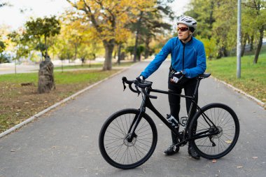 Gözlüklü ve koruyucu bisiklet kasklı bir sporcu bisiklet yolundaki parkta duruyor. Spor ve açık hava eğlencesi..