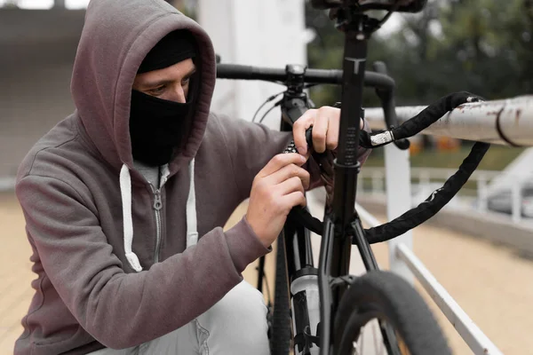 Biały złodziej w kominiarce i kapturze łamie zamek na rowerze na ulicy w ciągu dnia. Zamknięta twarz i hakowanie. Kradzież rowerów. — Zdjęcie stockowe