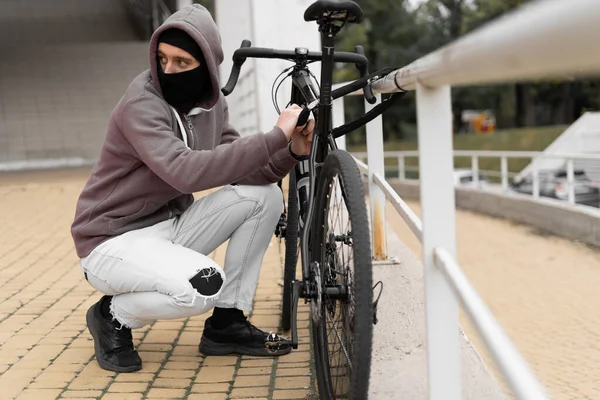 Ladrão caucasiano em balaclava e capuz quebra a fechadura em uma bicicleta na rua durante o dia. Cara fechada e pirataria. Roubar bicicletas. Imagens De Bancos De Imagens