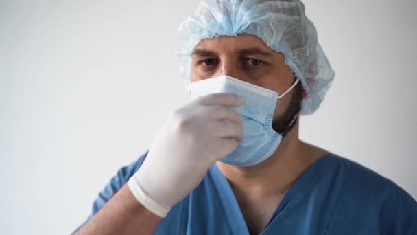 Trött manlig läkare kaukasiskt utseende på en vit bakgrund. Tar bort den medicinska masken från ansiktet. tar ett djupt andetag. Närbild av porträtt. — Stockvideo