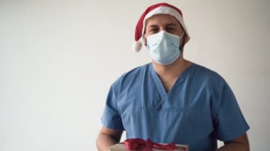 Tıbbi maske takmış ve Noel Baba şapkası takmış bir doktor. Elinde kırmızı bir fiyonk olan bir kutu tutuyor ve uzatıyor. Doktorlardan hediye konsepti