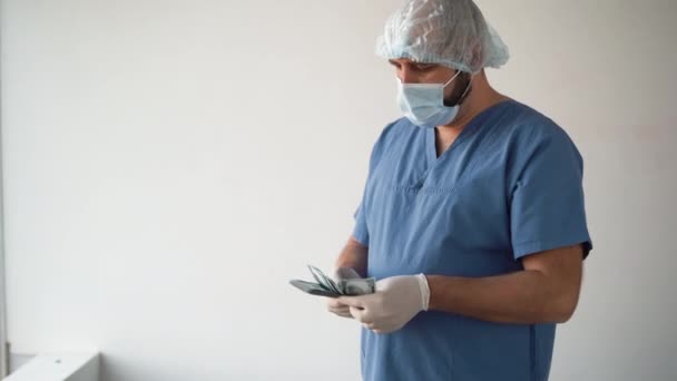 Bělošský lékař v lékařské masce a chirurgické čepici. stojí na bílém pozadí a počítá peníze hodí a odejde. 100 bankovek. — Stock video