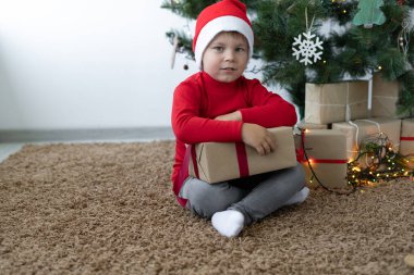 Beyaz çocuk, elinde kraft kağıdına sarılı bir kutu tutarak yerde oturuyor. Kafasında Noel Baba şapkası var. Boşluğu kopyala