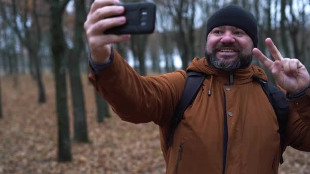 Homem de aparência de outono na floresta vestida com um chapéu preto e uma jaqueta marrom. Tira uma selfie ao telefone. Conceito da forma de um blogueiro na natureza — Vídeo de Stock