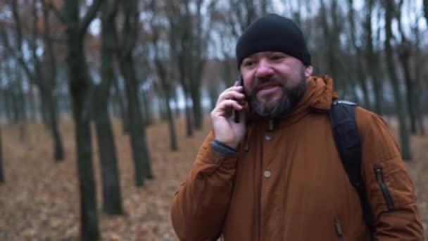 Homem de aparência de outono na floresta vestida com um chapéu preto e uma jaqueta marrom. Faz um telefonema e comunica — Vídeo de Stock