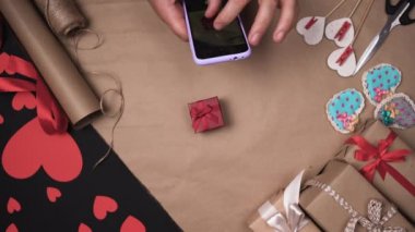 Erkek eller bir telefon tutuyor ve küçük kırmızı bir kutunun fotoğraflarını çekiyor. Paket kağıdı ve kırmızı kalpler. Sevgililer Günü hediye konsepti